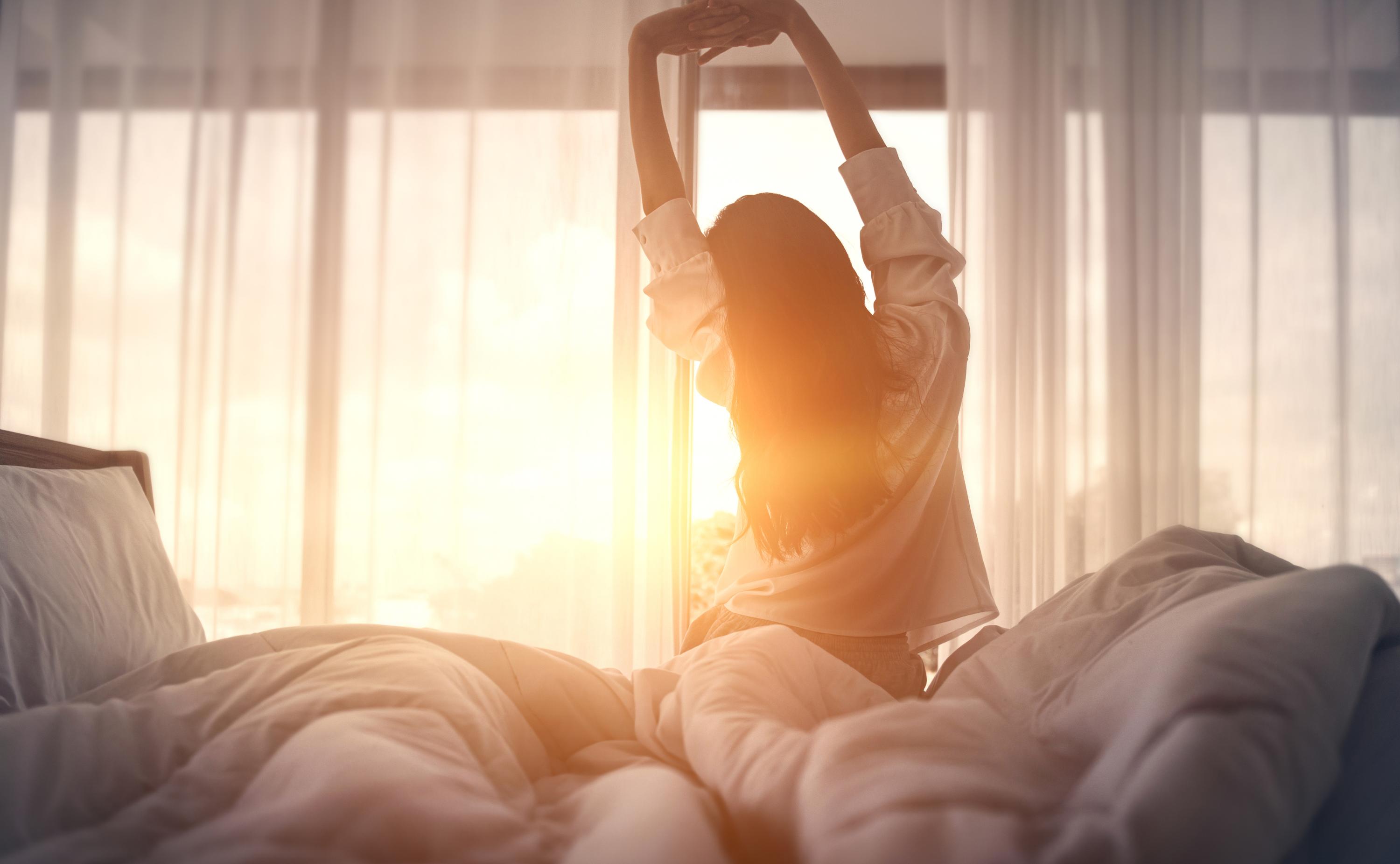 A woman sitting on her bed, stretching her arms in front of a window with sunlight beaming in