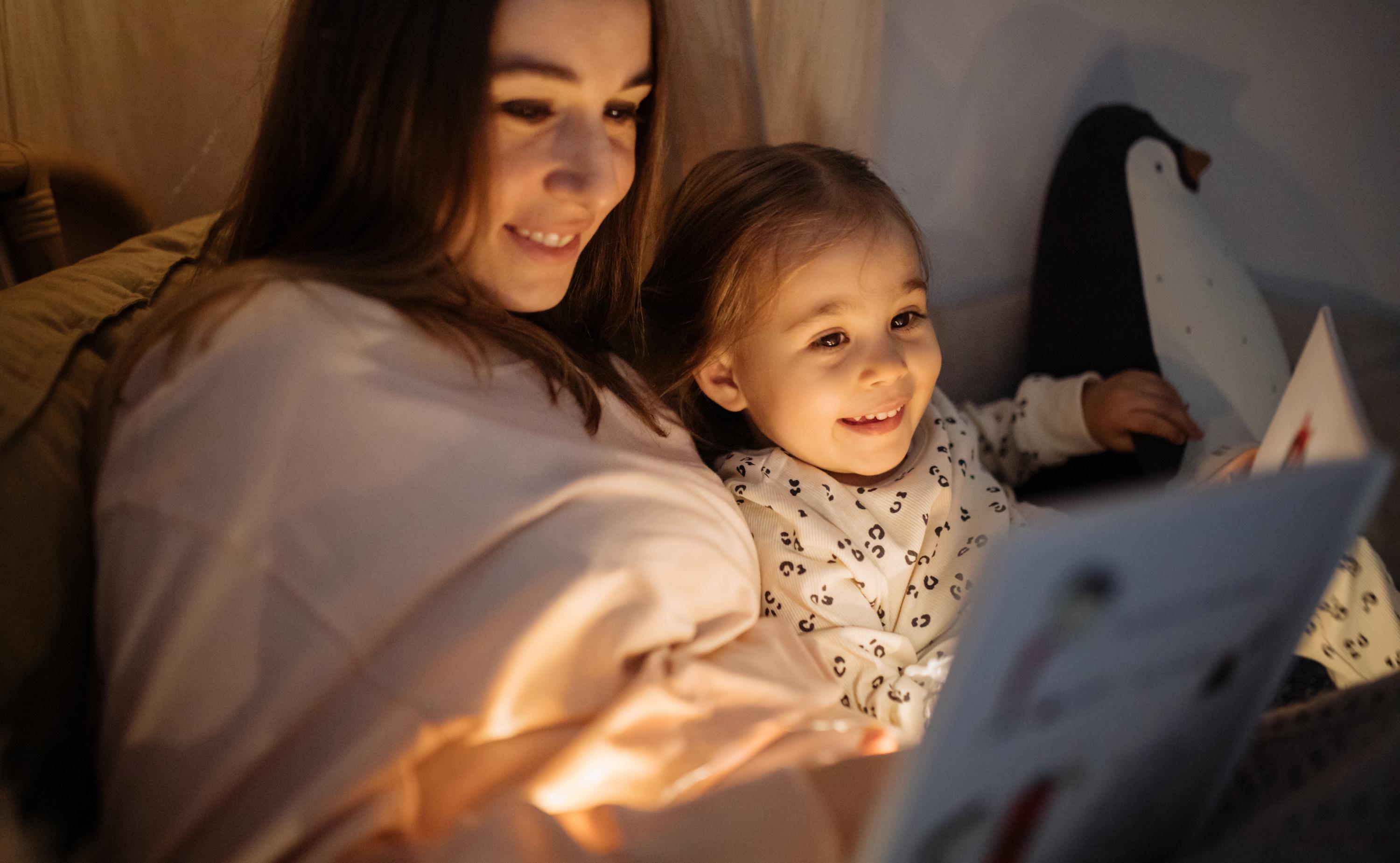 A woman lounging in a warmly dim lit space, reading to a child nestled in her arms.
