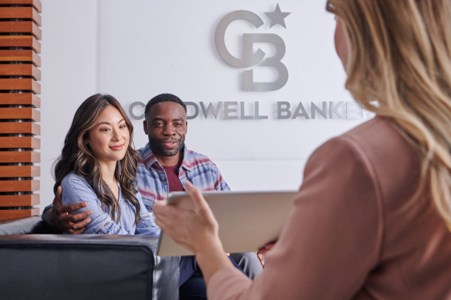 A young couple sitting down and facing a real estate agent who is explaining paperwork to them. Coldwell Banker logo is on the wall behind them.